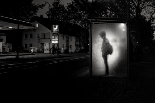 Black and white photo of a boy standing at a bus stop.